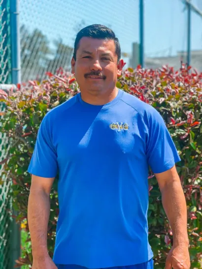 Man in blue shirt smiles, standing outdoors in front of green bushes.