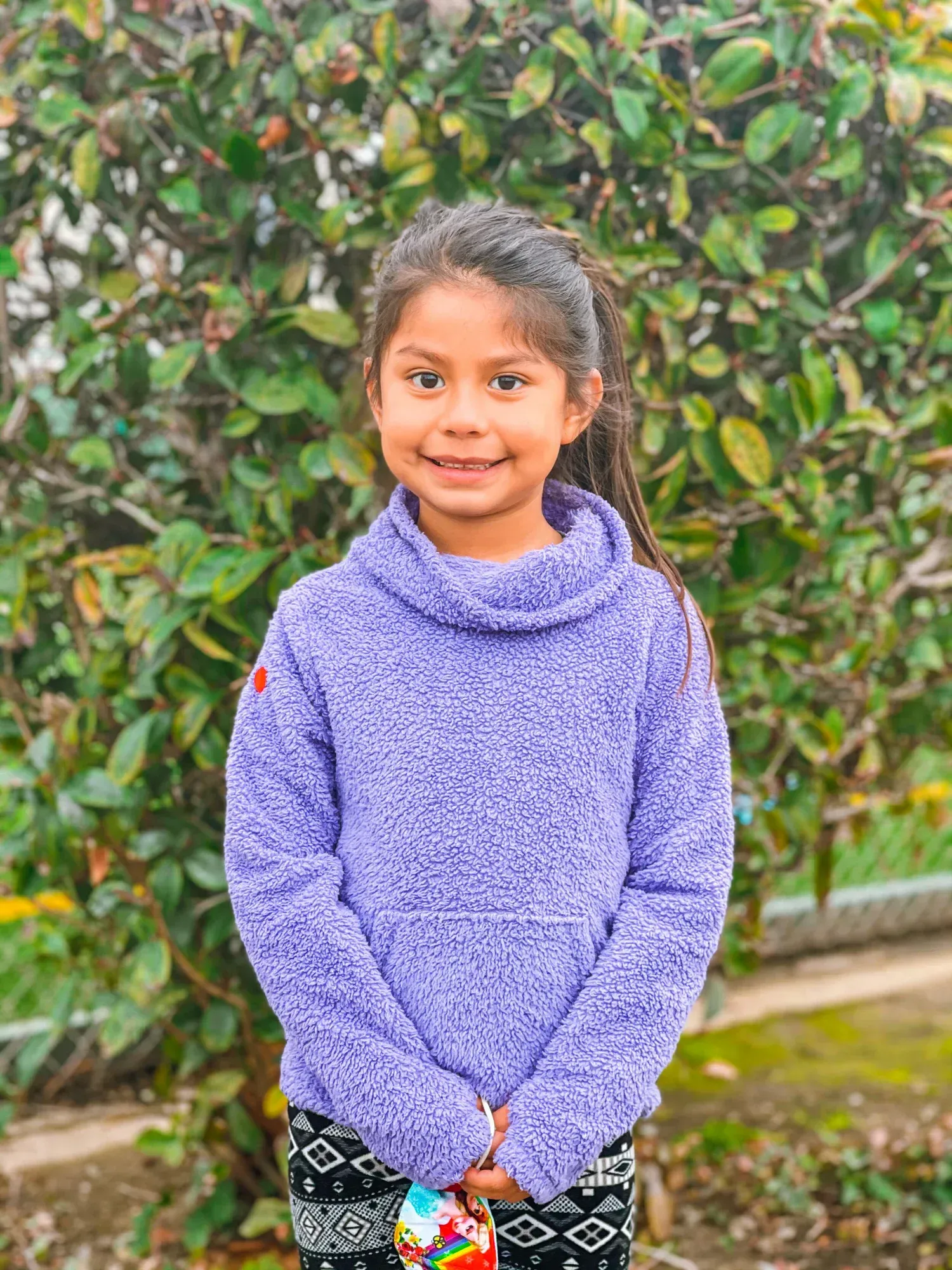 Girl in purple fleece sweatshirt smiles, standing in front of green shrubbery.