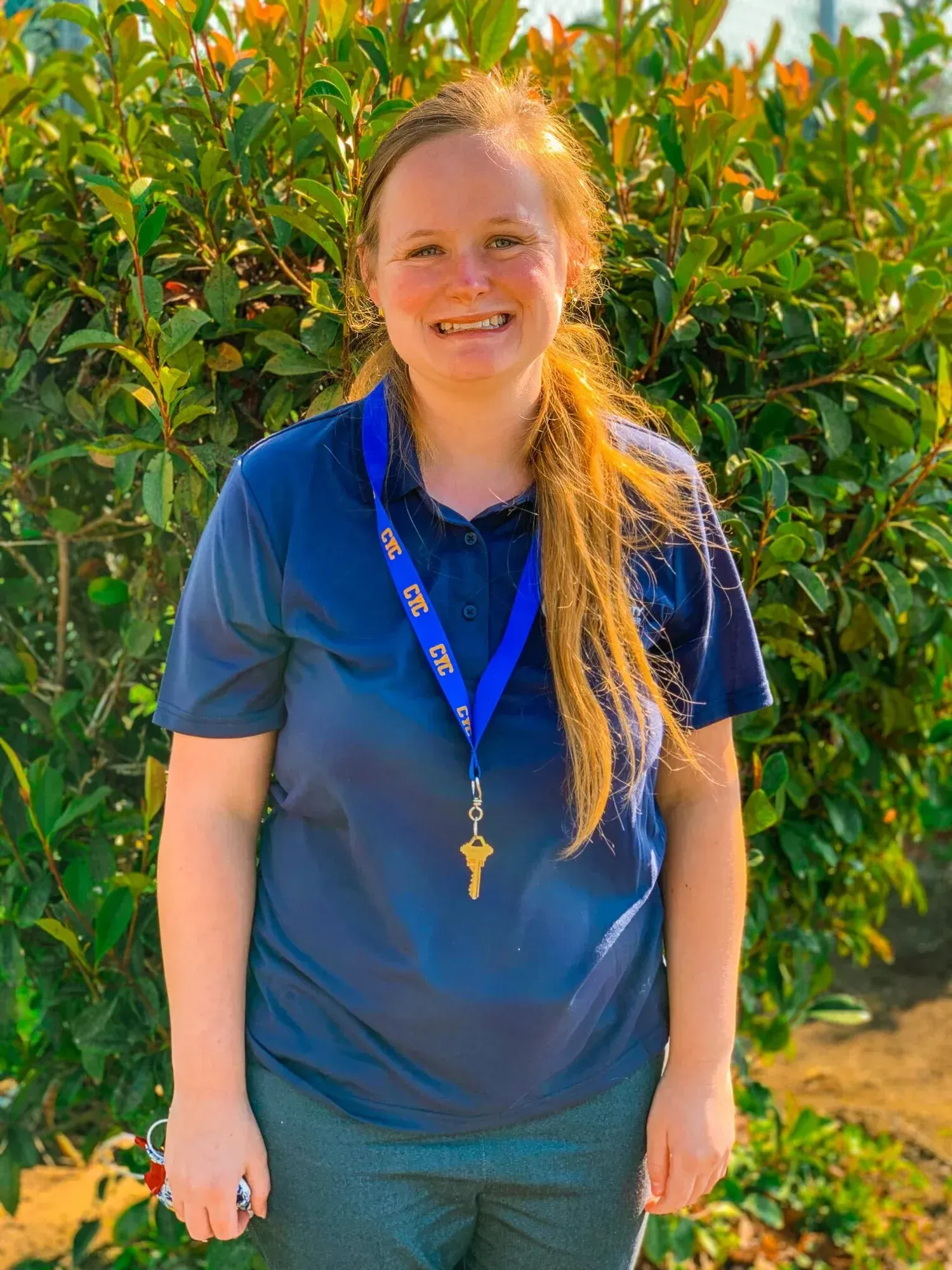 Woman in a blue shirt with a lanyard smiles in front of green foliage.