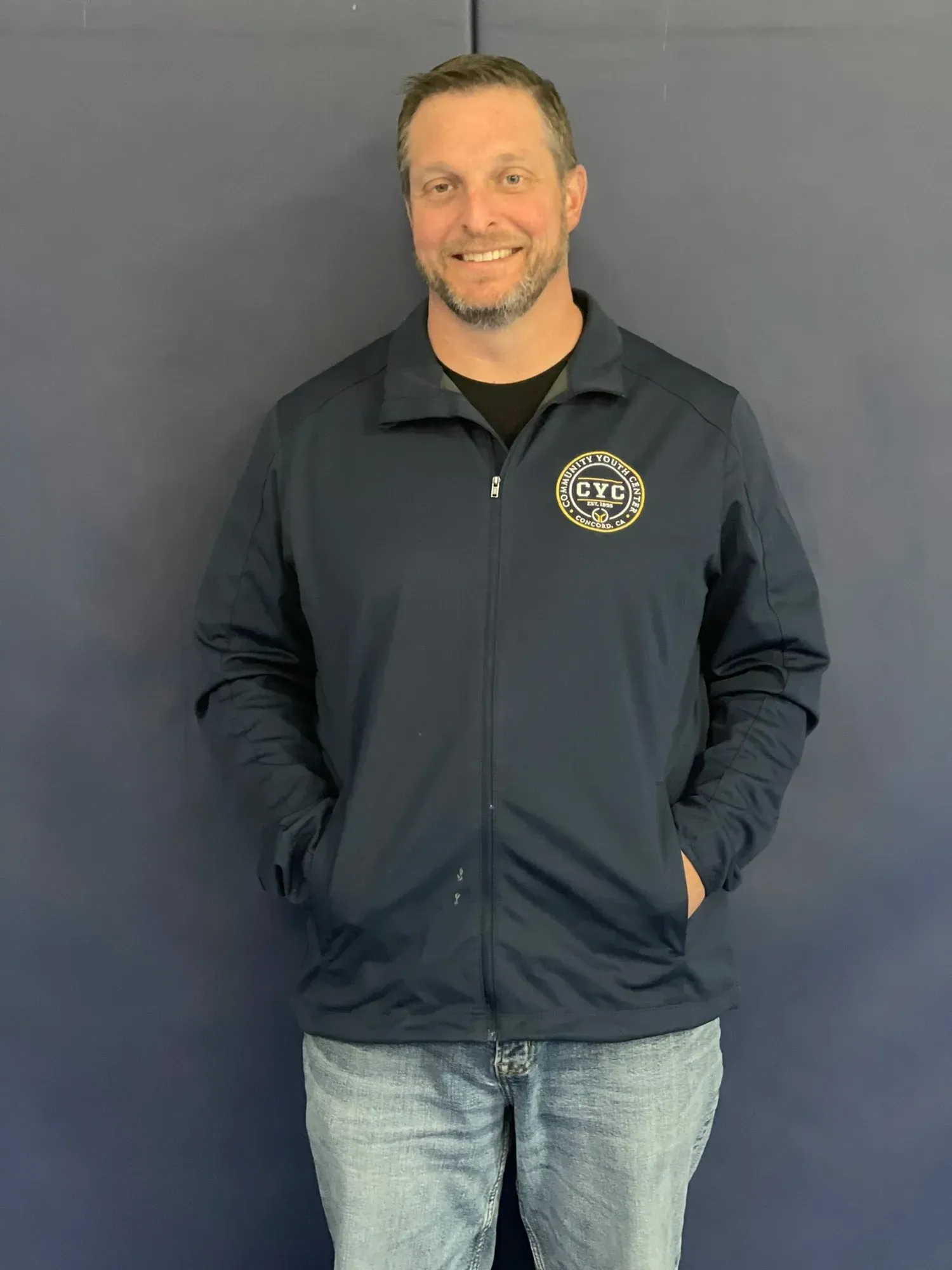 Man in blue jacket and jeans smiles, hands in pockets, against a blue wall.