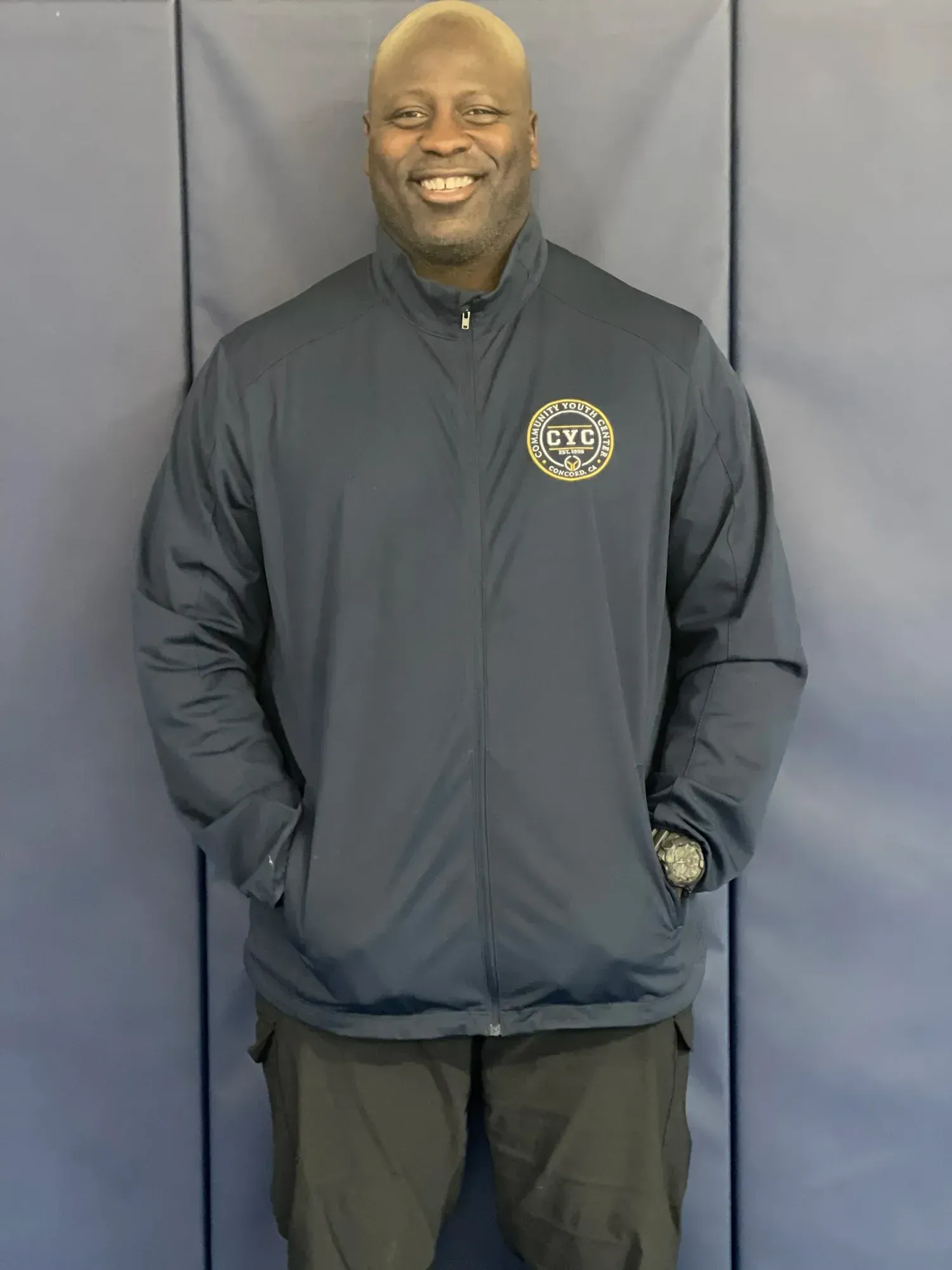 Man in navy jacket with emblem smiles, hands in pockets, against blue textured wall.