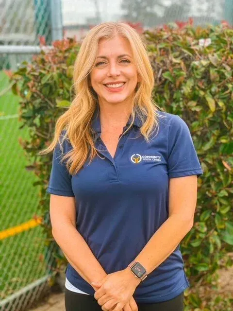 Woman in navy polo shirt smiles, arms crossed, in front of greenery and sports field.