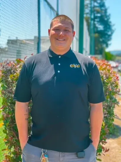 Man in black polo shirt with hands in pockets stands outside near bushes.