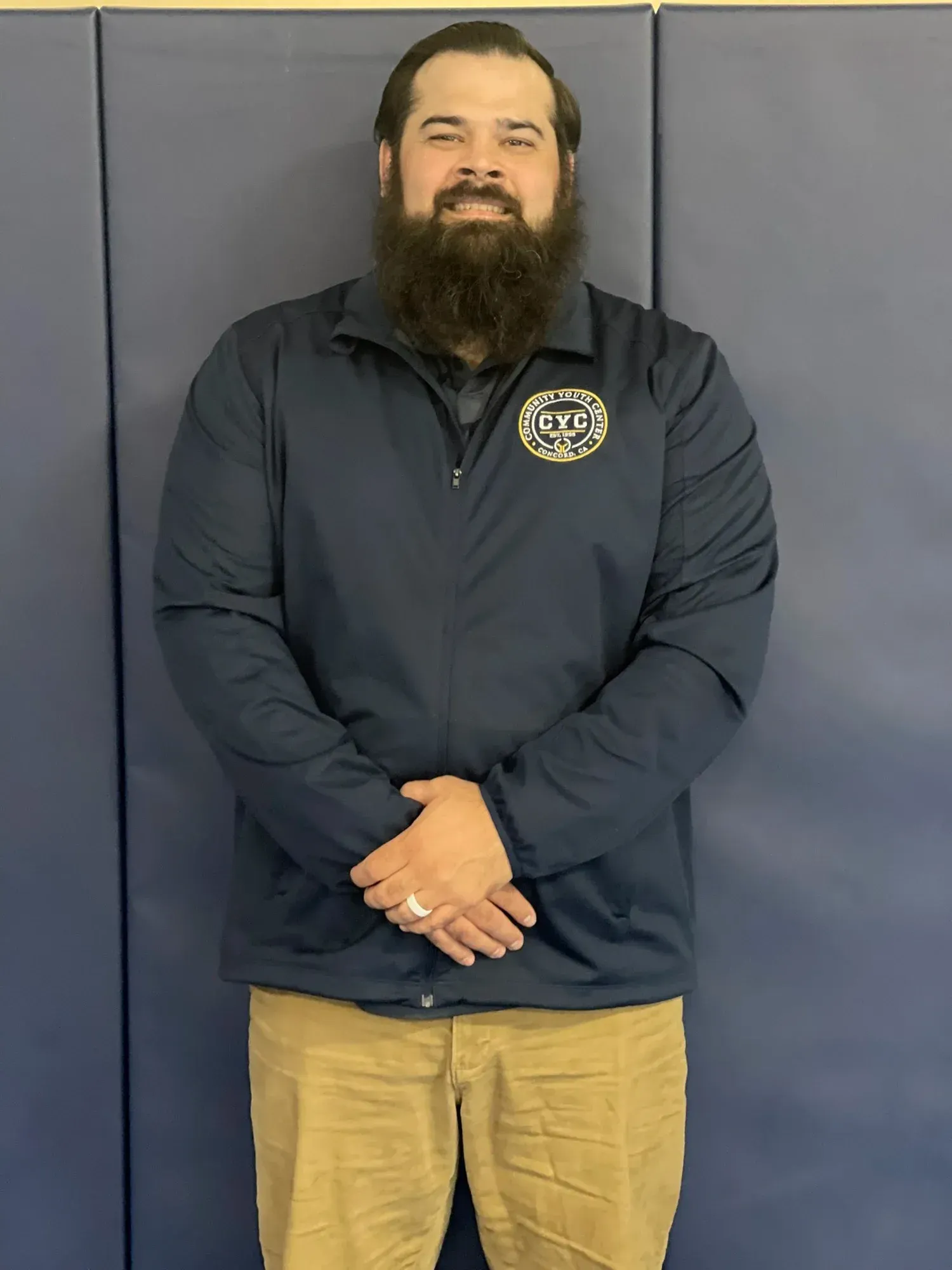 Man with a full beard, wearing a navy jacket and khakis, standing in front of a blue wall.