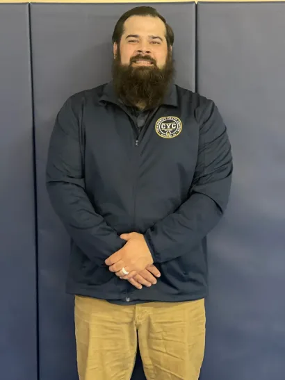 Man with a full beard, wearing a navy jacket and khakis, standing in front of a blue wall.