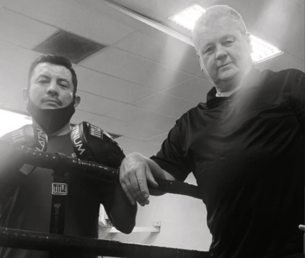 Two men in a boxing ring. One wearing a face mask leans on the ropes, the other, with a black shirt, looks at the camera.