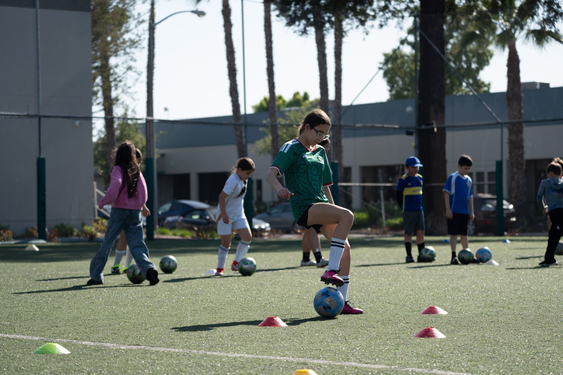 Children playing soccer on a green field with cones. One child in orange runs with the ball.