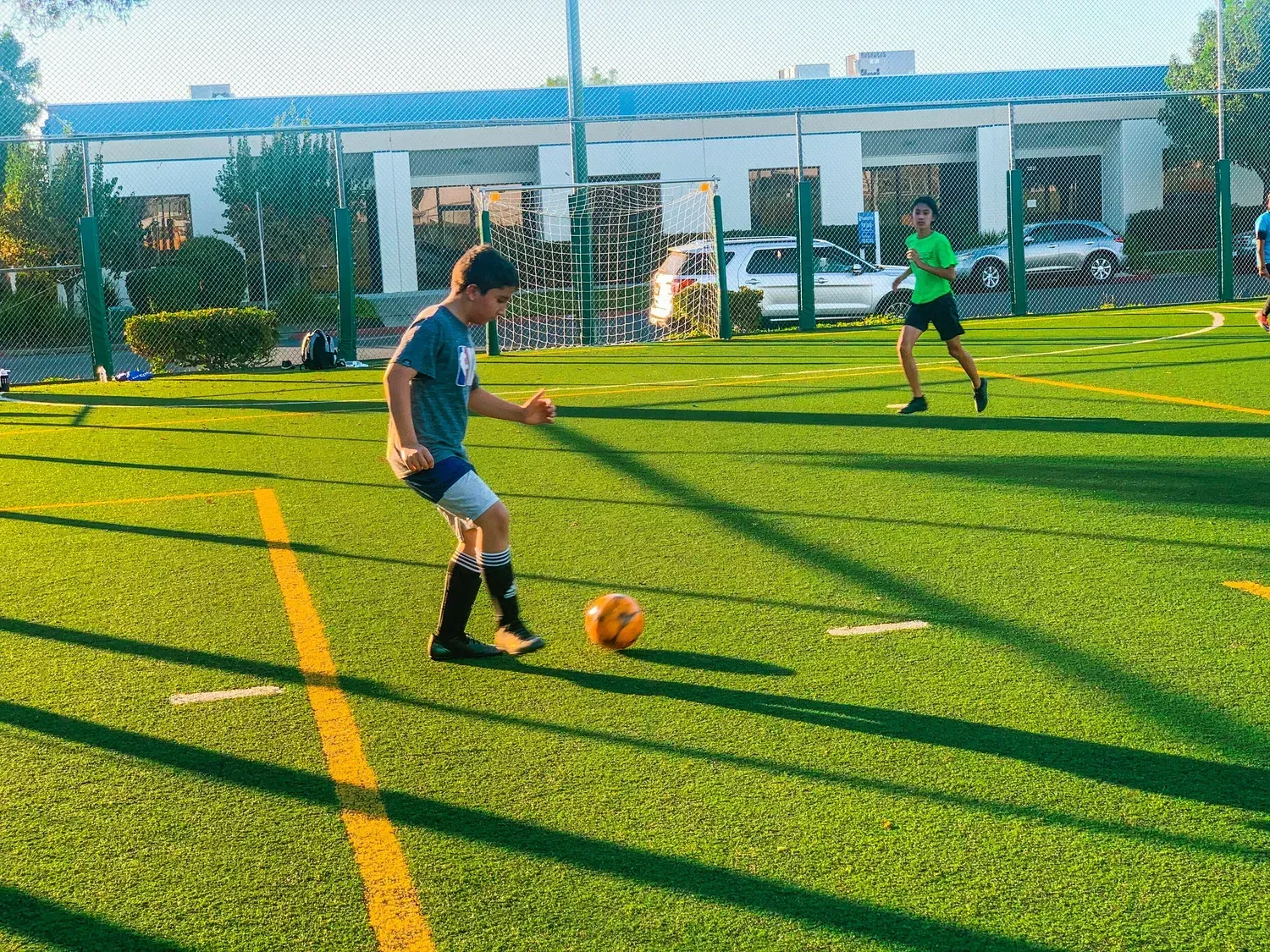 Boy in a gray shirt dribbles a soccer ball on a green field, another player runs in the background near a building.