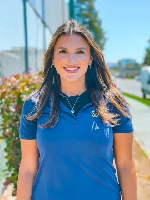 Woman smiling, wearing blue polo shirt, necklace, earrings, outdoors near greenery and street.