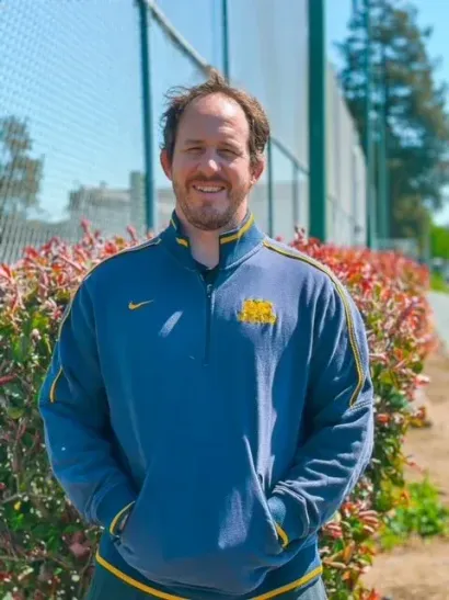 Man in blue sweatshirt with gold accents, standing outdoors near bushes and a chain-link fence, smiling.