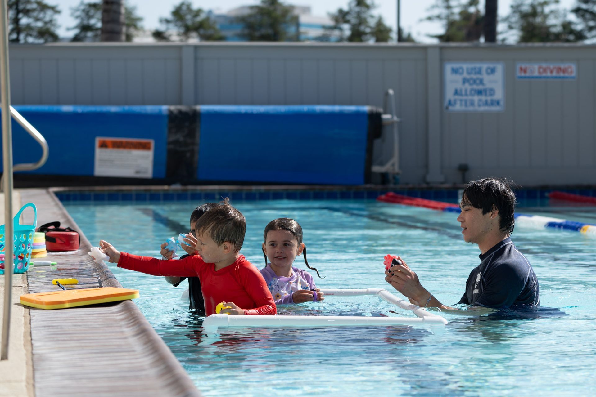 Children and an instructor in a pool, reaching for toys near the edge