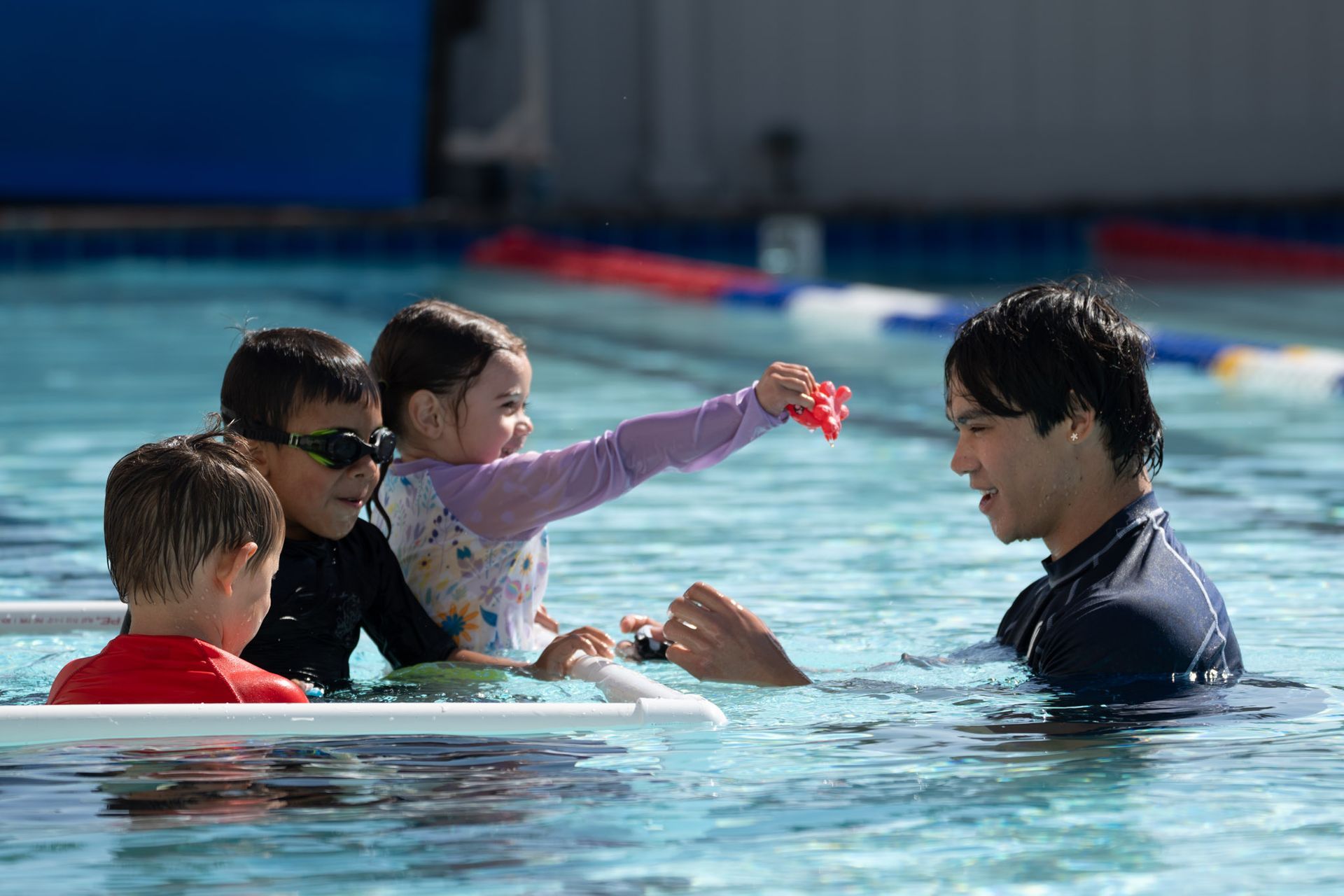 Swim instructor and three children in a pool, one child holding out a red toy to the instructor.