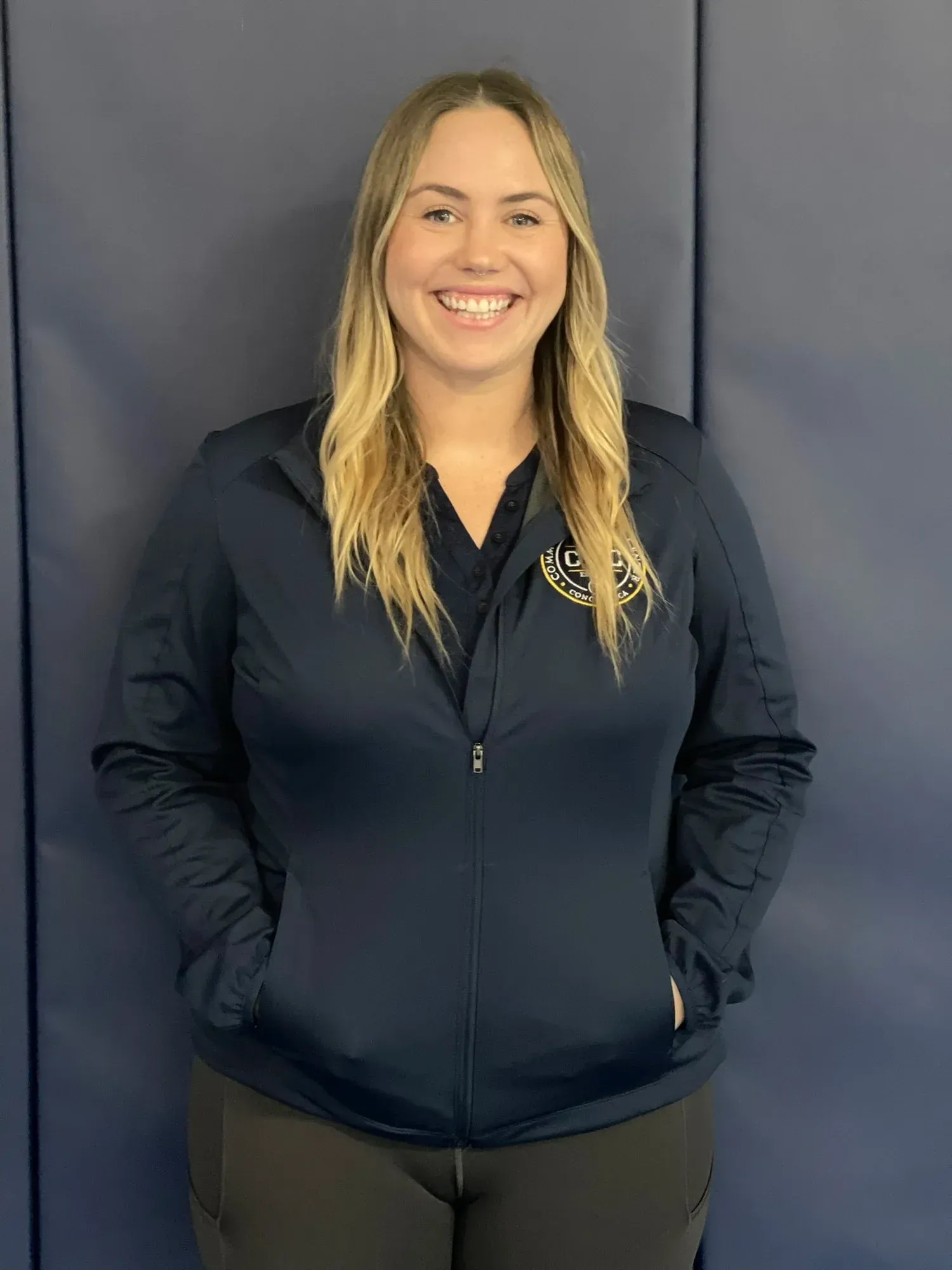 Woman in a navy jacket smiling with hands in pockets; standing in front of a blue wall.