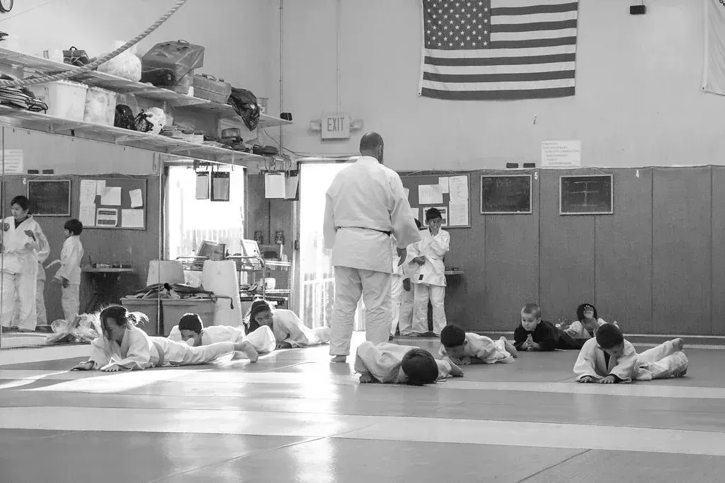 Judo class. Students in white uniforms on a mat, instructor facing them under American flag.