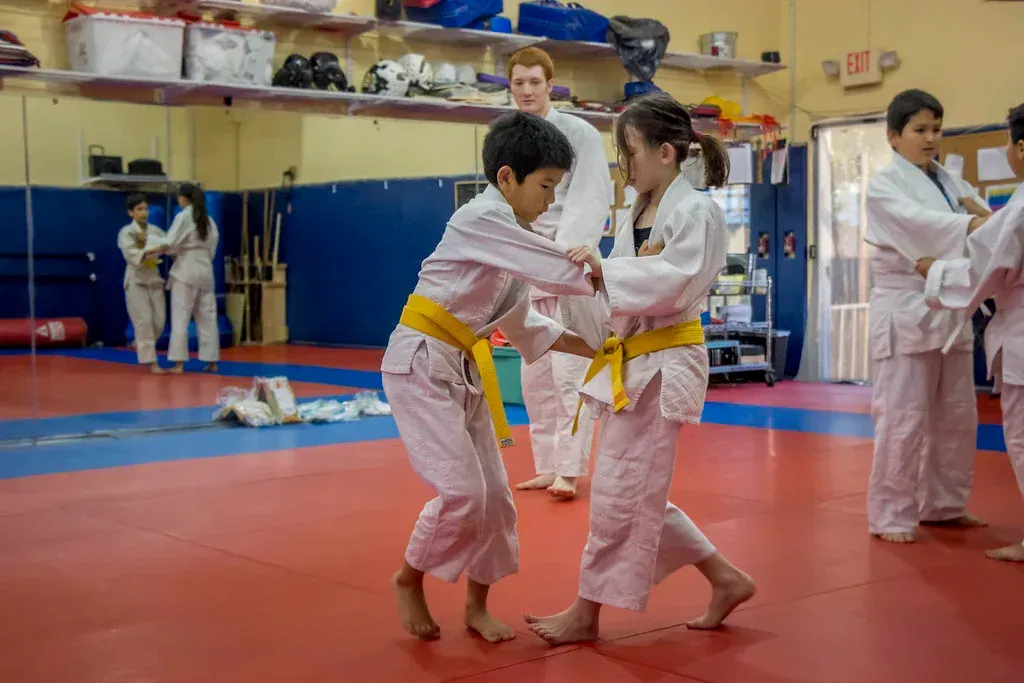 Children in white uniforms with yellow belts practice Judo on a red mat, with an instructor in the background.
