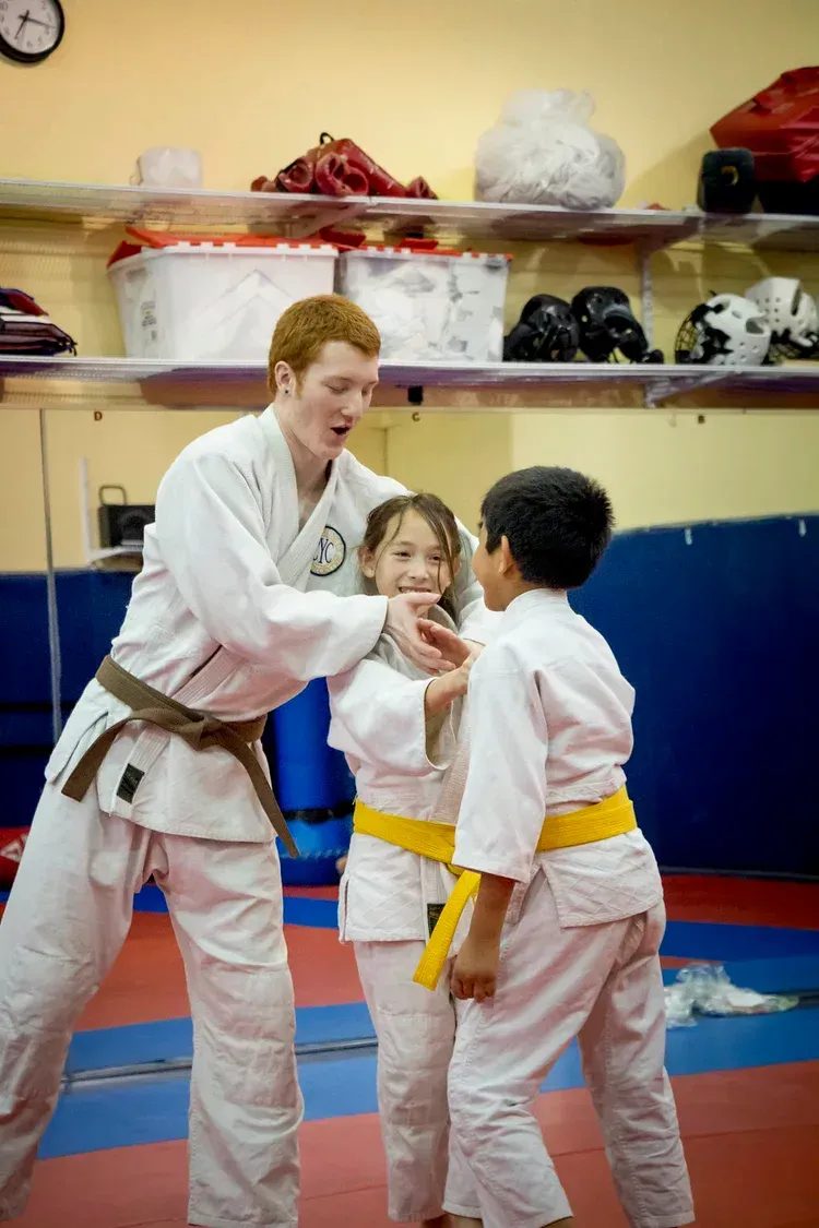 Martial arts instructor in a white uniform demonstrating a technique to two children in yellow belts.