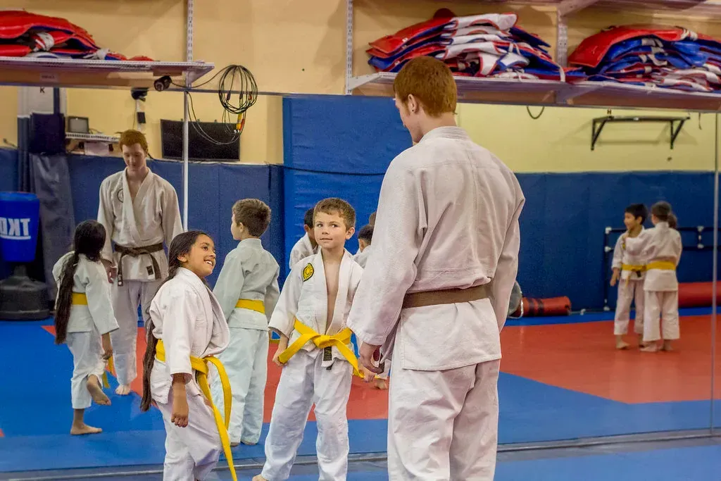 Kids in karate uniforms with a trainer in a dojo, blue mats, mirrored wall.