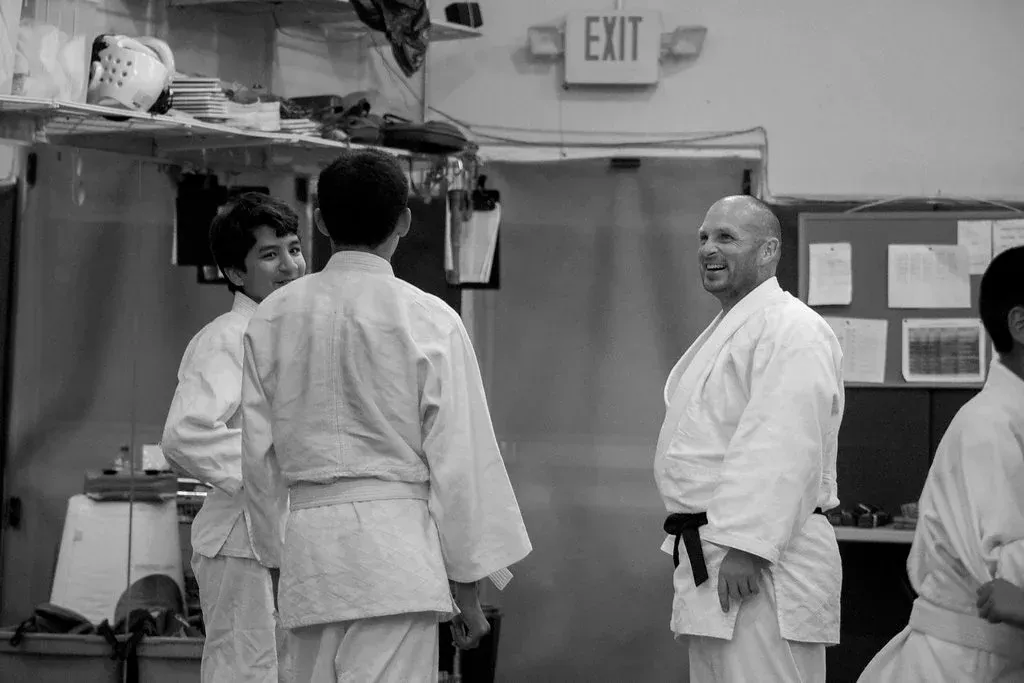 People in white martial arts uniforms in a dojo. A smiling man with a black belt stands near two others.
