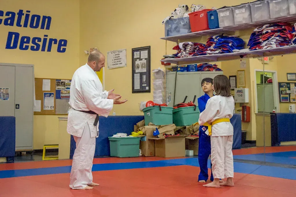 Man in white judo uniform instructing two children in a dojo.