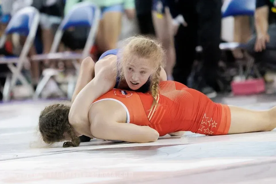 Two wrestlers grappling on a mat, one in blue on top of one in orange, in an arena.