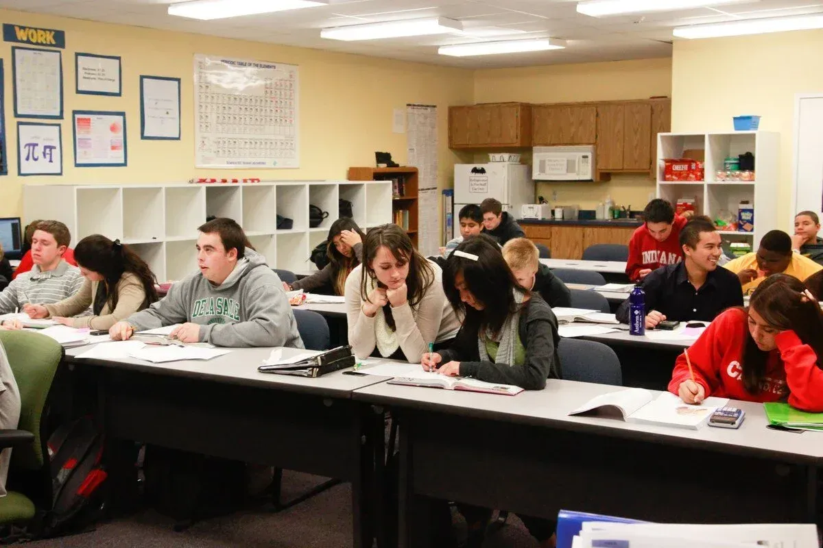 Students seated at desks in a classroom, studying or writing. Some are focused, others look on.