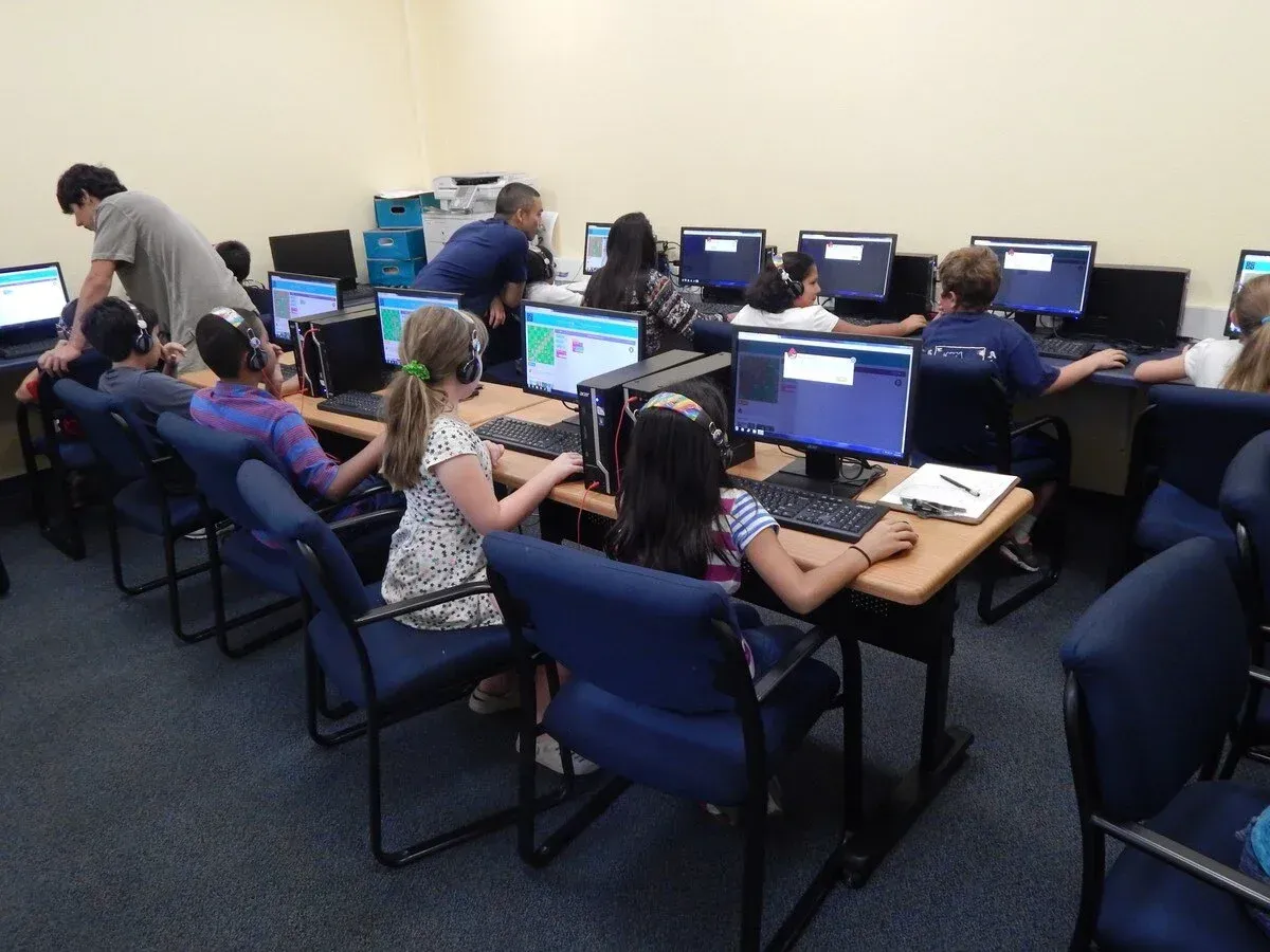 Children in a computer lab, some wearing headsets, learning with monitors. Two adults assist students.