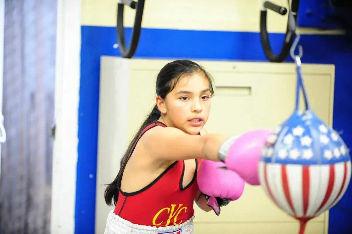 Girl boxing in a gym, punching a red, white, and blue speed bag, wearing pink gloves and red leotard.