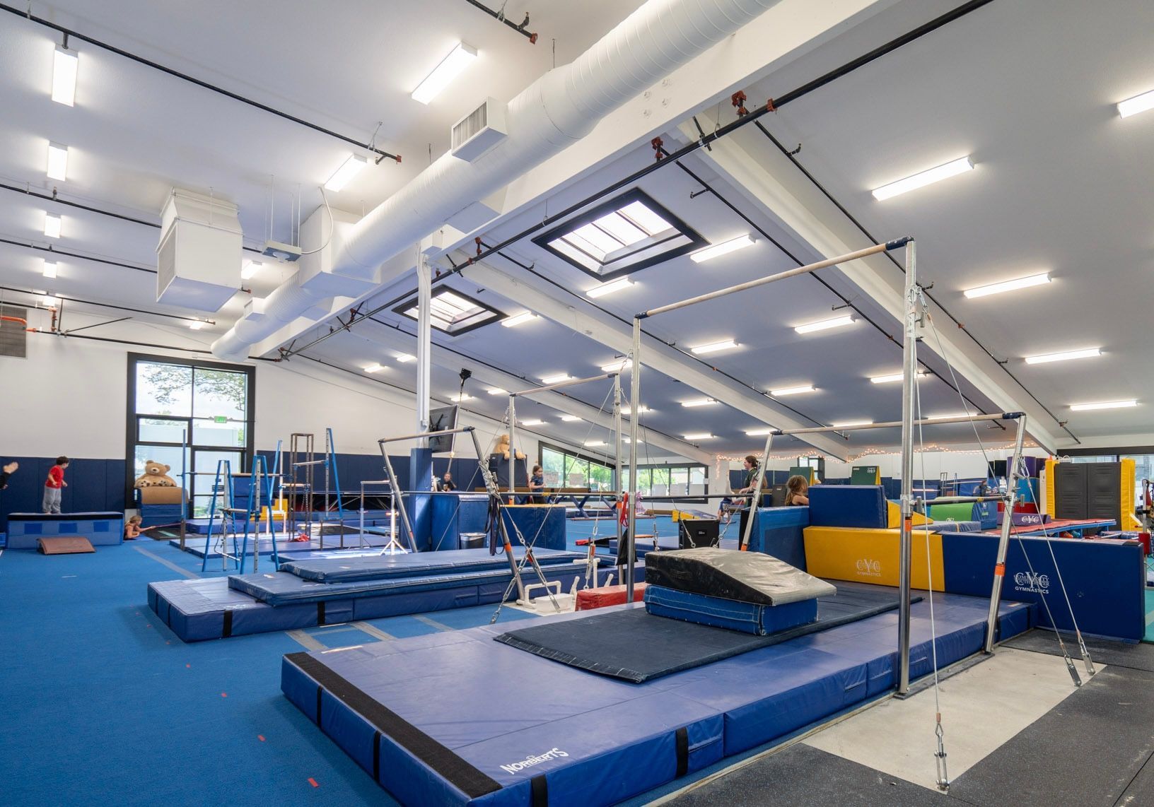Indoor gymnastics training gym with blue mats, trampolines, and metal frames under bright lights
