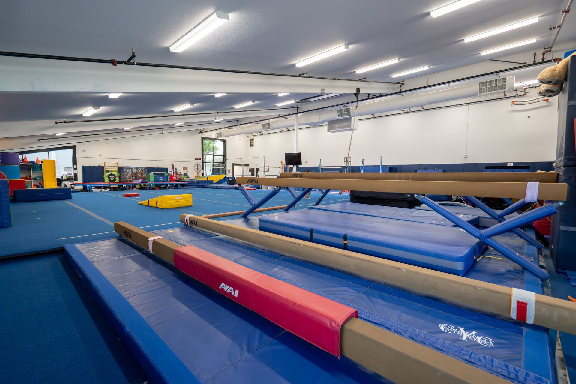 Indoor gymnastics gym with blue mats, balance beams, and training equipment under bright lights