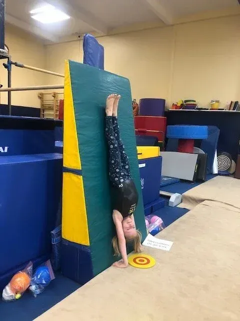 Girl doing a handstand against a padded wall in a gymnastics gym, with a target on the floor.