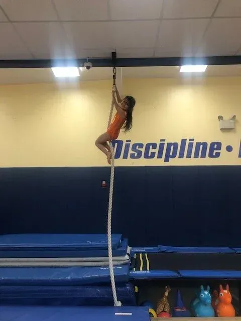 Girl in orange leotard climbing a rope in a gym, blue and yellow walls visible.