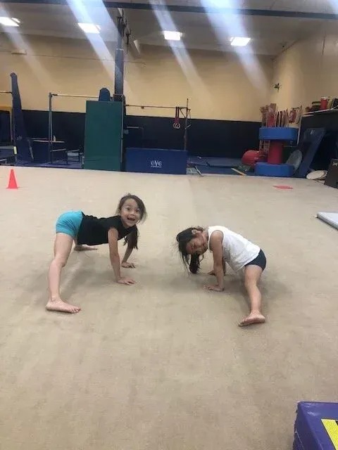 Two girls smiling, doing gymnastics stretch on gym floor.