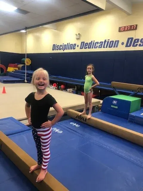 Two girls on balance beams in a gym; one smiling, wearing leggings, the other in a leotard.