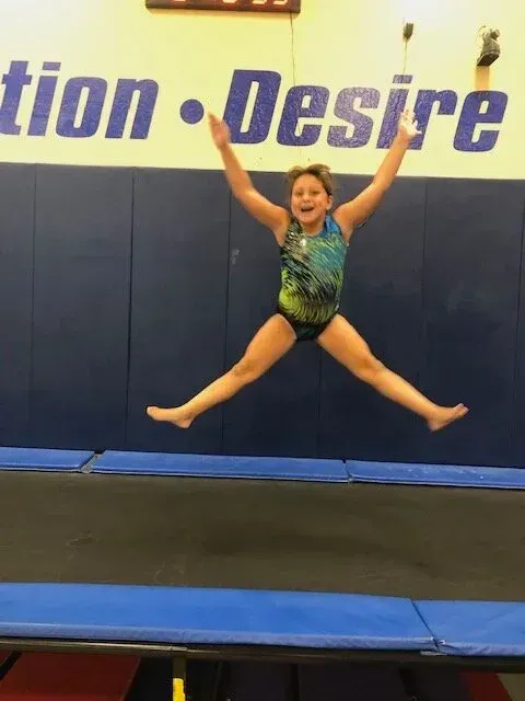 Gymnast jumping high in the air on a trampoline with arms raised, legs split. Blue and yellow gym.
