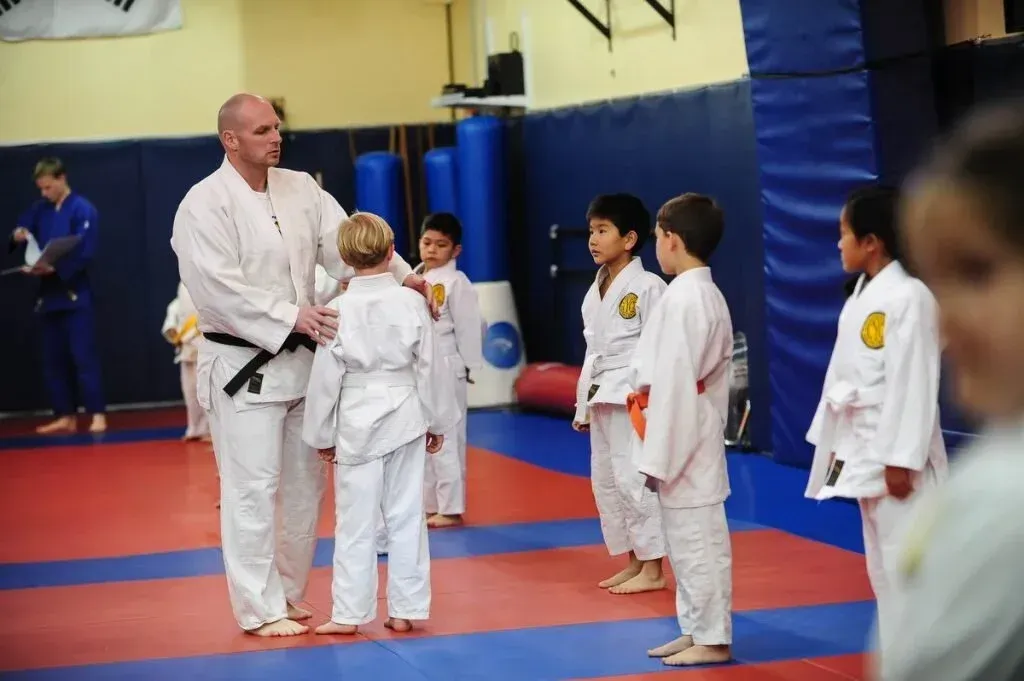 Martial arts instructor with students, teaching in a gym with blue mats, wearing white uniforms.