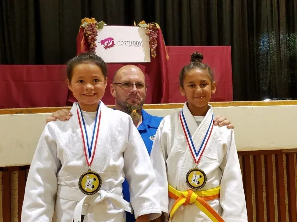 Two children in judo uniforms with medals posing with a man in front of a stage.