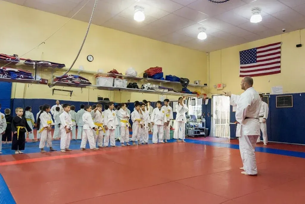 Karate class: Instructor facing students in white uniforms, yellow belts, standing on red mat, American flag in background.