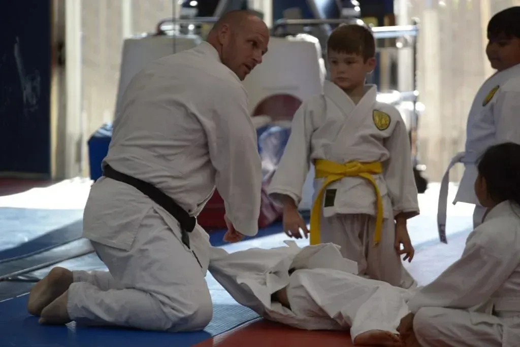 Instructor demonstrates a technique to young students on a mat; one child is lying down.