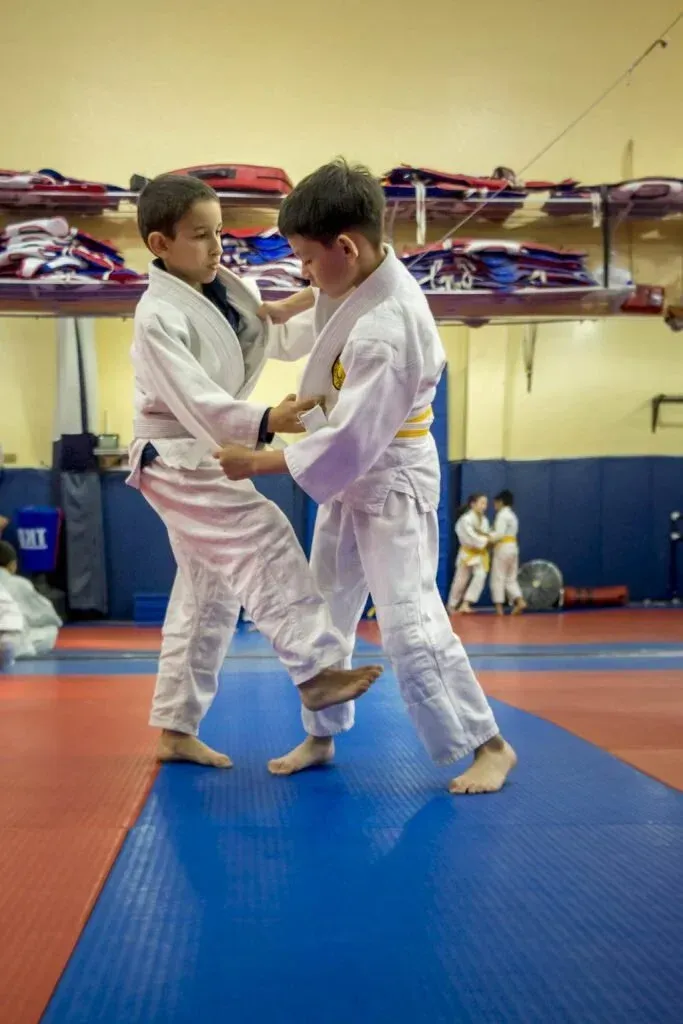Two young judo students in white uniforms practice a foot sweep technique on a blue mat.