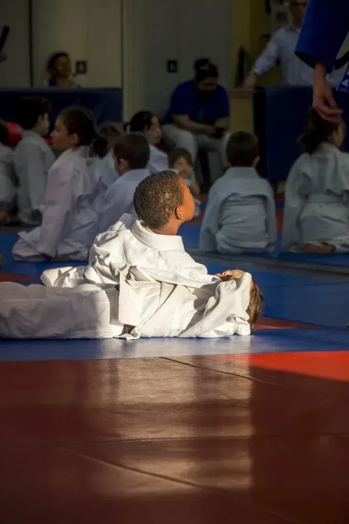 Boy in white judo uniform sits on a red and blue mat, looking up, with other children in the background.