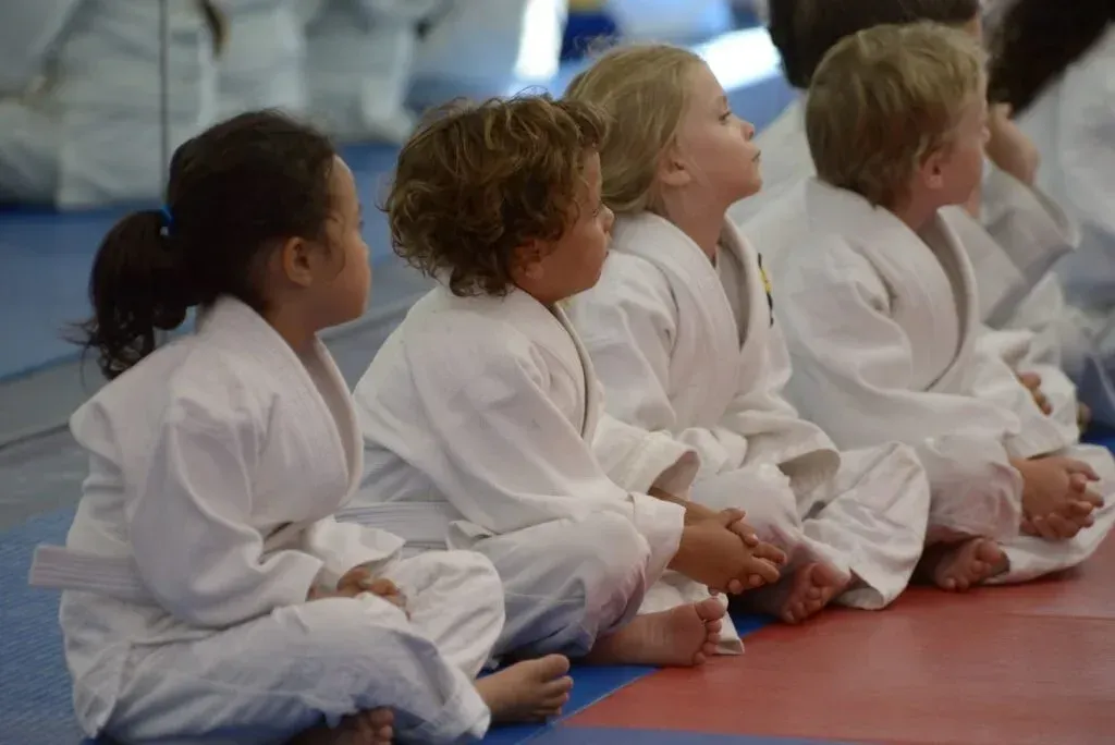 Children in white martial arts uniforms seated on a mat, looking ahead.