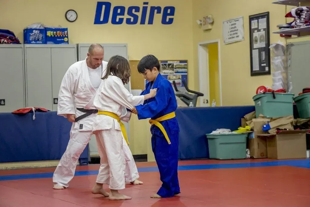 Judo class: Instructor in white gi watches two children in gis practice a technique on red mat.