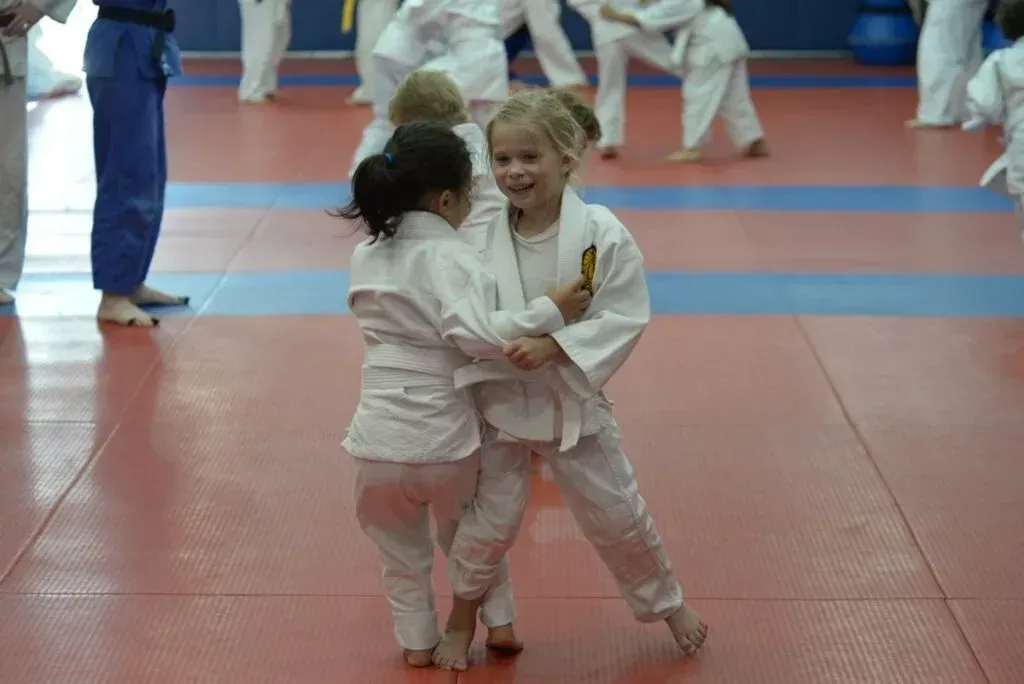 Two children in white judo uniforms facing each other on a red and blue mat, smiling and practicing.