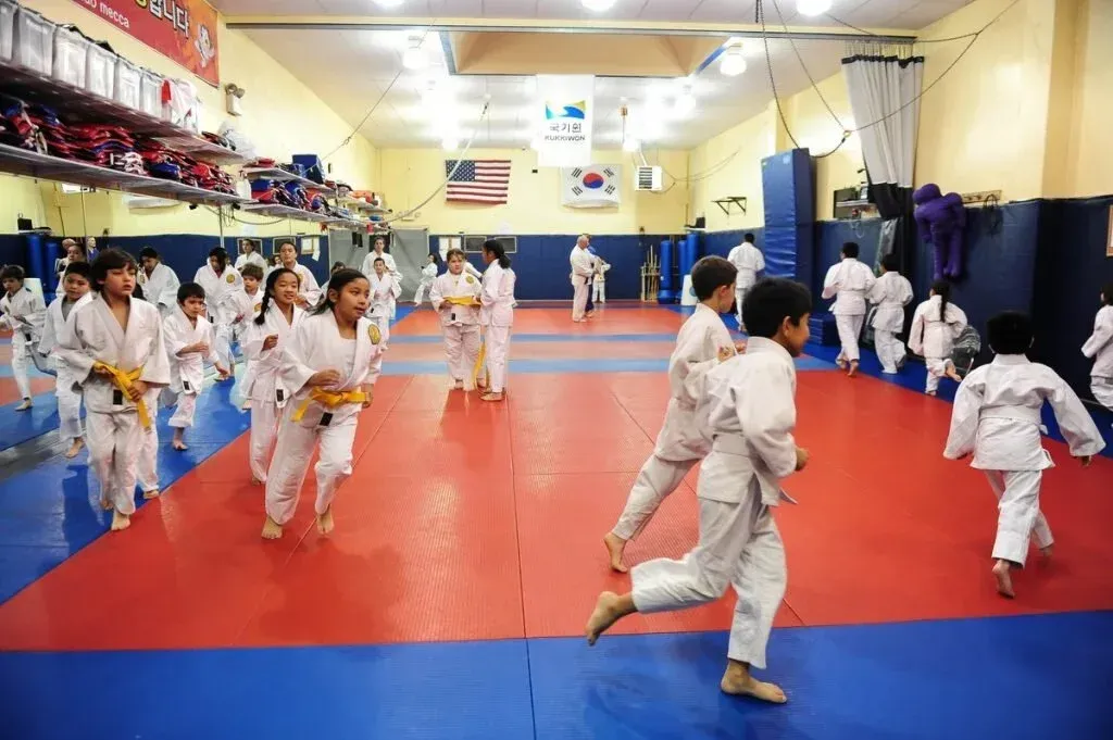 Children in white martial arts uniforms running on red and blue mats in a gym.