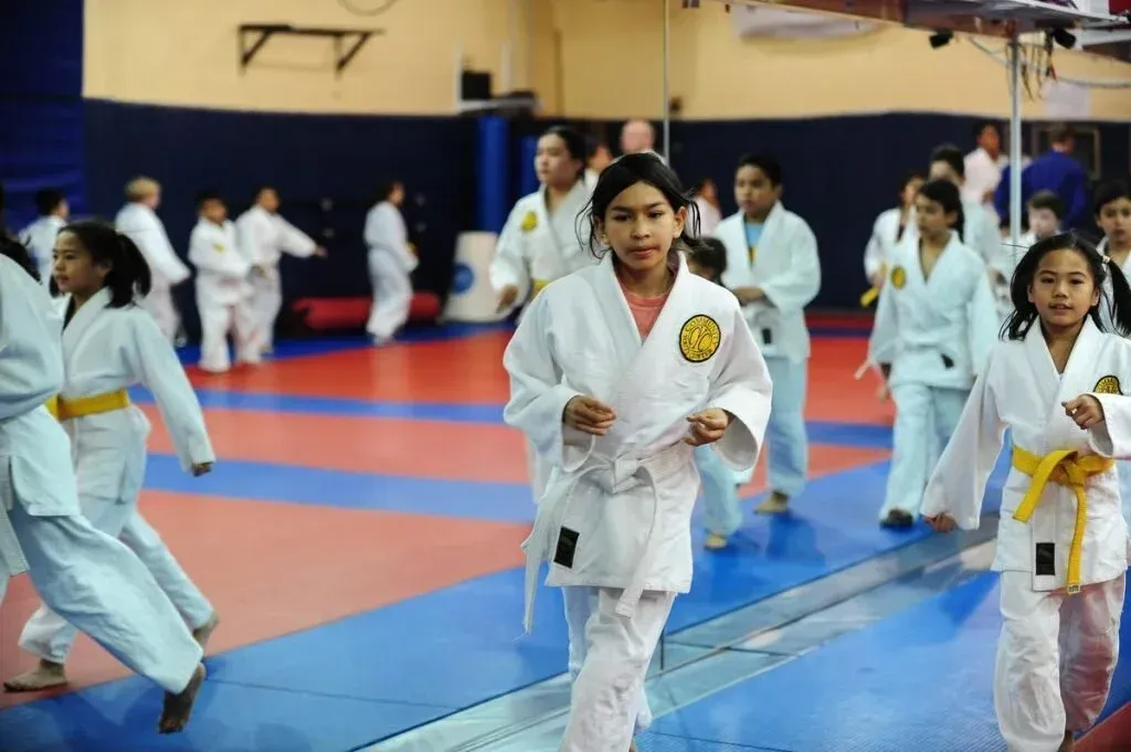 Children in white judo uniforms with yellow belts on a blue and red mat.
