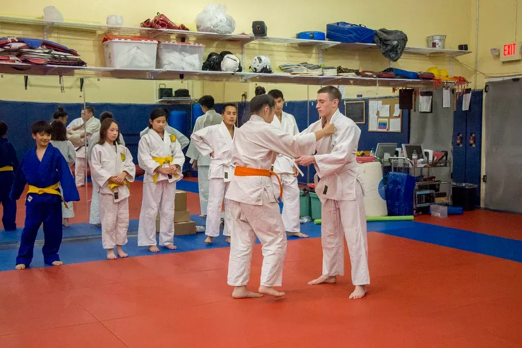 Judo class in progress, students in white judo uniforms, orange and yellow belts, training on a red mat.