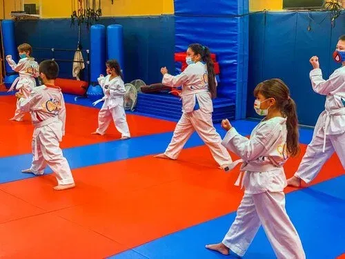 Children in karate uniforms practice martial arts in a gym with red and blue mats.