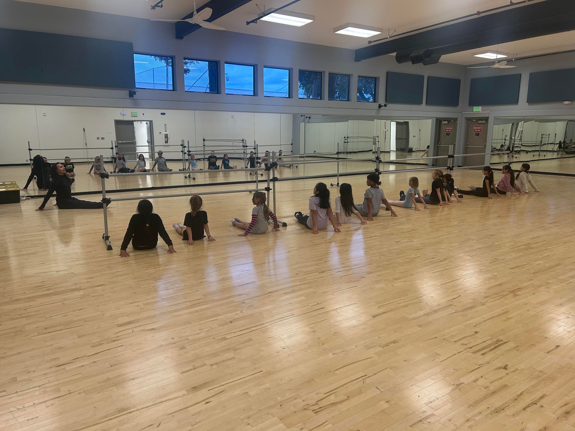 Young dancers practice ballet at a barre in a studio, arms outstretched.