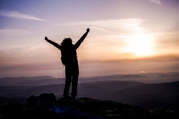Silhouette of person with arms raised, atop a mountain, against sunset.