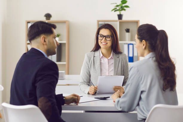 Woman in glasses smiles, speaking to a couple at a table. Office setting with documents.